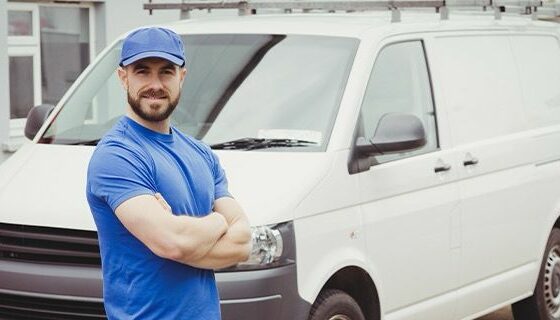 na imagem, vemos um homem de braços cruzados em frente a uma van no estacionamento de uma empresa de receptivos turísticos