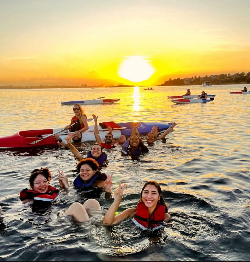 na imagem, vemos um grupo de pessoas dentro do mar usando colete salva-vidas na cor vermelha, sorridentes, olhando para a câmera.