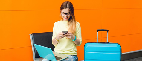 na imagem, vemos uma moça loira sentada em um banco ao lado de uma mala azul e com um notebook em seu colo da mesma cor. a imagem serve para ilustrar o texto sobre a importãncia de investir em reservas online durante eventos regionais.