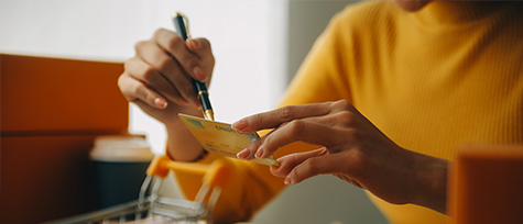 na imagem, vemos uma pessoa segurando um cartão de crédito em frente a um computador. Ao lado, um carrinho de compras em miniatura.